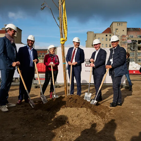 Sechs Personen mit Helmen pflanzen gemeinsam einen Baum auf einer Baustelle.