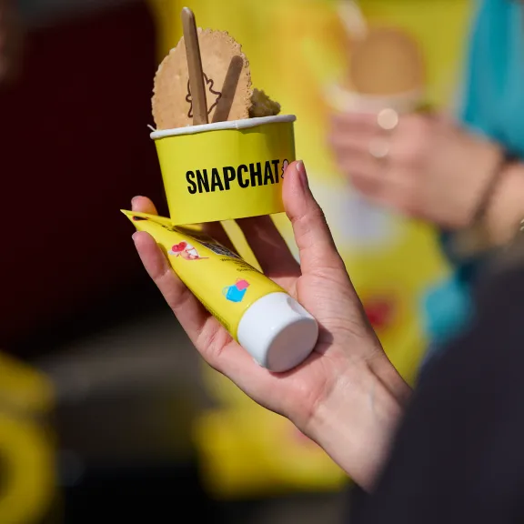 Hand hält Snapchat-Eisbecher mit Schokoladeis und gelber Tube, unscharfer Hintergrund.