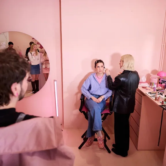 Visagistin schminkt Frau in rosafarbenem Schminkstudio, Spiegel reflektiert weitere Personen.
