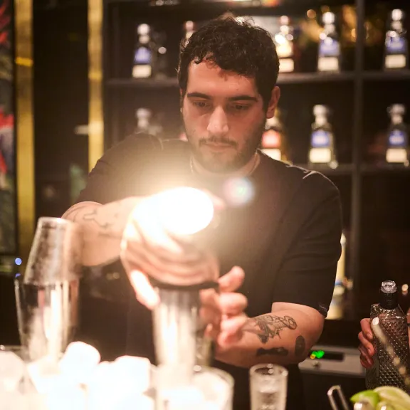 Barkeeper mixt einen Drink an einer beleuchteten Bar, Flaschen im Hintergrund.