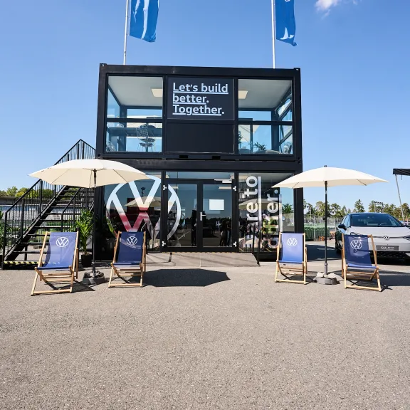 Modernes, zweistöckiges Gebäude mit VW-Logo, Liegestühlen und Sonnenschirmen vor blauem Himmel.
