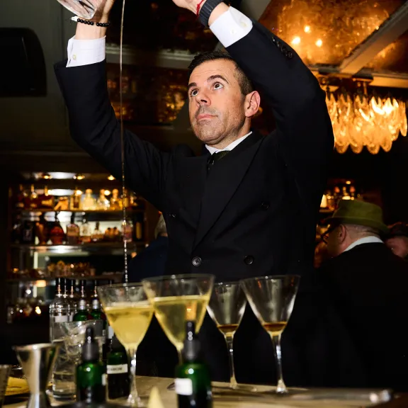 Barkeeper gießt Cocktail in Gläser, Bar mit Flaschen im Hintergrund, festliche Stimmung.