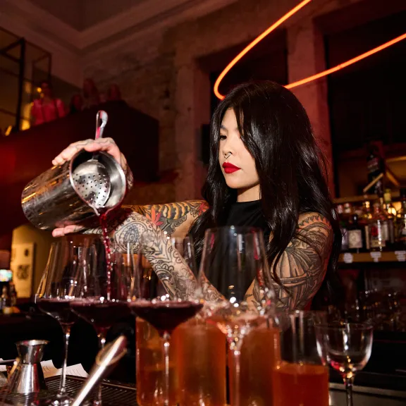 Frau hinter Bar gießt roten Cocktail in Gläser, stimmungsvolles Licht.