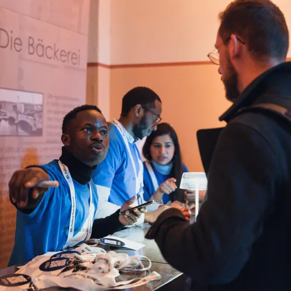 Ein Mann am Empfangstisch spricht mit einem Besucher, mehrere Personen im Hintergrund.