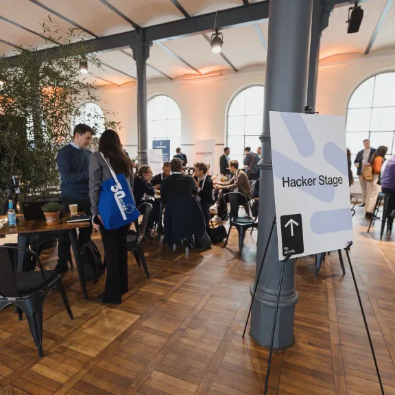 Menschen unterhalten sich an Stehtischen in hellem Raum, großes Fenster im Hintergrund.
