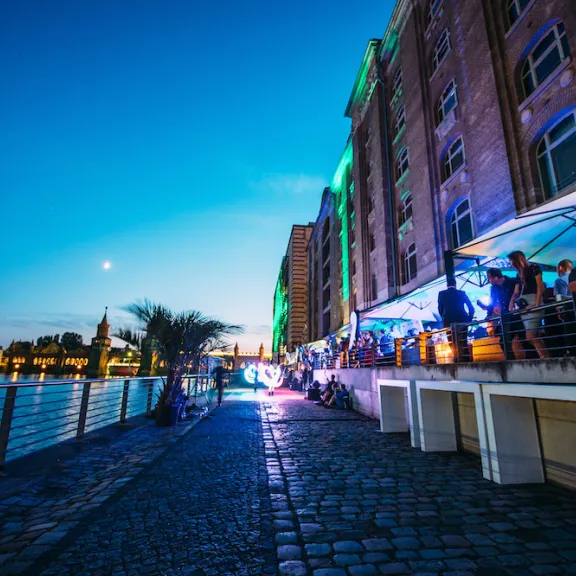 Menschen sitzen abends in einem beleuchteten Restaurant am Flussufer.