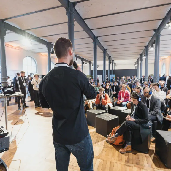 Mann spricht bei einer Konferenz zu einem sitzenden Publikum in modernem, hellen Raum.
