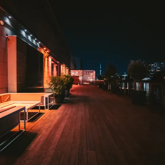Beleuchtete Terrasse mit Holzboden und Bänken nachts, Stadtlichter im Hintergrund.