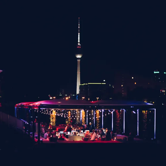 Beleuchtete Dachterrasse mit Menschen, im Hintergrund der Berliner Fernsehturm bei Nacht.