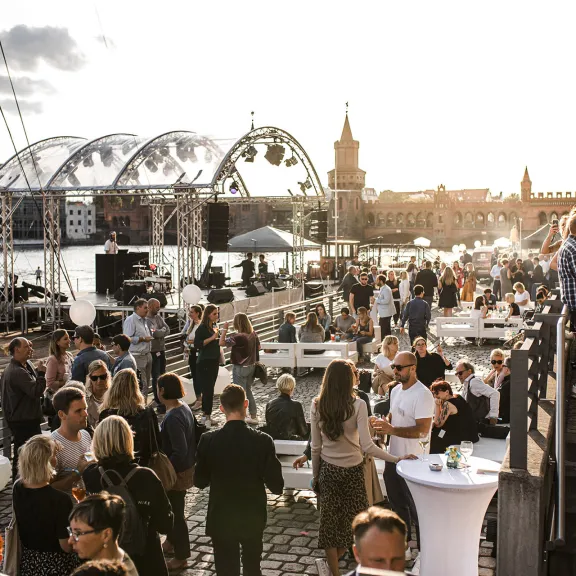 Menschen feiern draußen an einem sonnigen Tag am Wasser mit Brücken im Hintergrund.