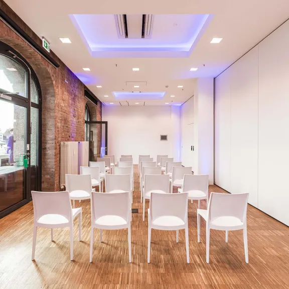 Heller Seminarraum mit weißen Stühlen in Reihen, Holzboden und Backsteinwand.