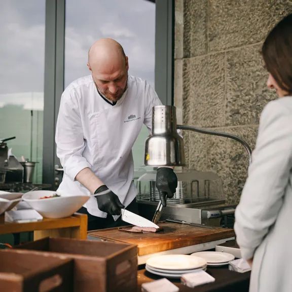 Koch bereitet Speisen an einer Theke vor, Frau steht gegenüber und schaut zu.