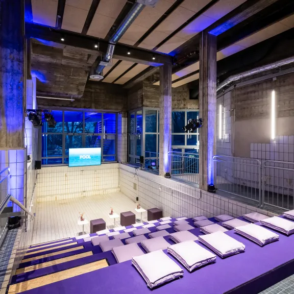 Moderner Veranstaltungsraum mit lila Sitzpolstern, Beton, Fliesen und blauer Beleuchtung.