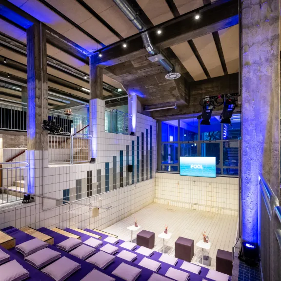 Modernes Auditorium mit gestuften Sitzreihen, lila Kissen, Scheinwerfern und Leinwand.