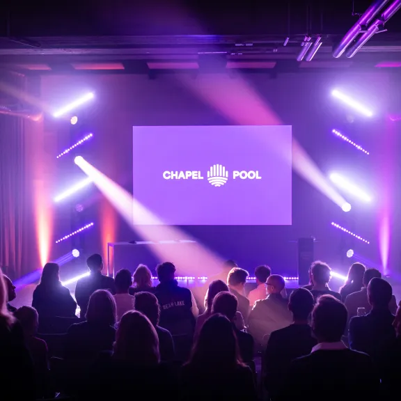 Publikum sitzt vor einer beleuchteten Bühne mit lila Licht und großem Bildschirm.