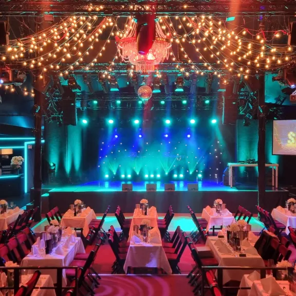 Festlich dekorierter Veranstaltungssaal mit gedeckten Tischen und bunter Bühnenbeleuchtung.
