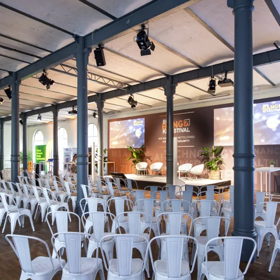 Leerer Veranstaltungsraum mit weißen Stühlen und Bühne, auf der Leinwände projiziert werden.