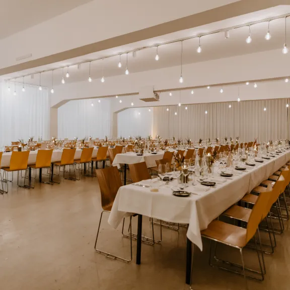 Festlich gedeckter Saal mit langen Tafeln, Holzstühlen und warmem Licht.