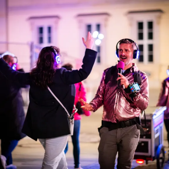 Moderator mit Mikrofon und Funkkopfhörern begleitet Teilnehmende einer Silent Disco Tour, die ebenfalls Kopfhörer tragen und im urbanen Außenraum interaktiv teilnehmen.