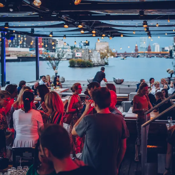 Menschen feiern am Abend auf einer Terrasse am Wasser, Lichterketten sorgen für Stimmung.