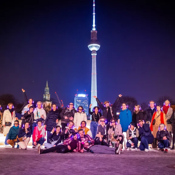 Gruppe von Teilnehmenden mit leuchtenden Funkkopfhörern posiert nachts vor dem Berliner Fernsehturm während einer Silent Disco Tour.