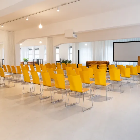 Heller Konferenzraum mit gelben Stühlen, Beamer und Leinwand.