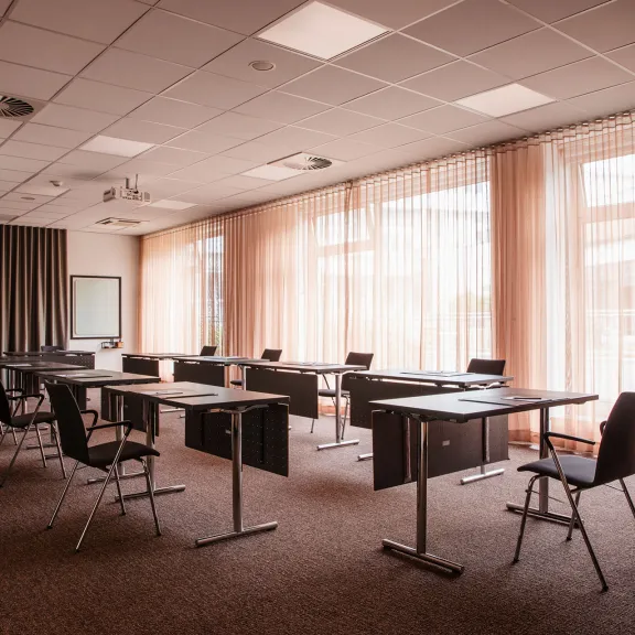 Heller Seminarraum mit Tischen, Stühlen und großen Fenstern mit Vorhängen.
