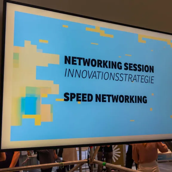 Monitor mit Schriftzug „Networking Session Innovationsstrategie Speed Networking“ vor Messestand.