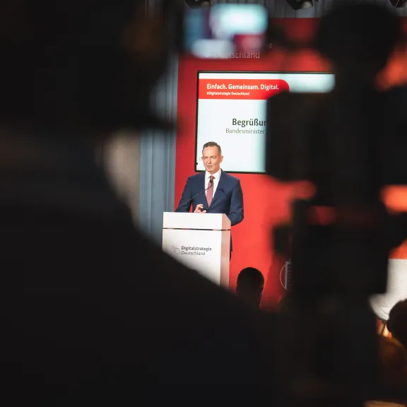 Mann im Anzug hält Rede auf Bühne, Kamera im Vordergrund, roter Hintergrund.