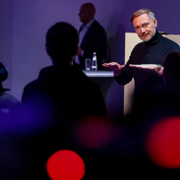 Mann spricht gestikulierend vor Publikum in dunklem Raum, Hintergrund unscharf.