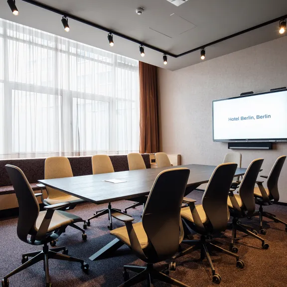 Moderner Konferenzraum mit Tisch, Stühlen und großem Bildschirm.