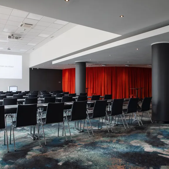 Leerer Konferenzraum mit schwarzen Stühlen, rotem Vorhang und Projektorleinwand.