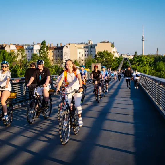 Menschen auf Fahrrädern und Inlineskates überqueren eine Brücke bei Sonnenschein.