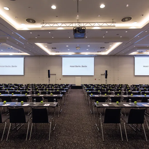 Großer, heller Konferenzraum mit vielen leeren Tischen und Stühlen, drei Leinwände vorne.
