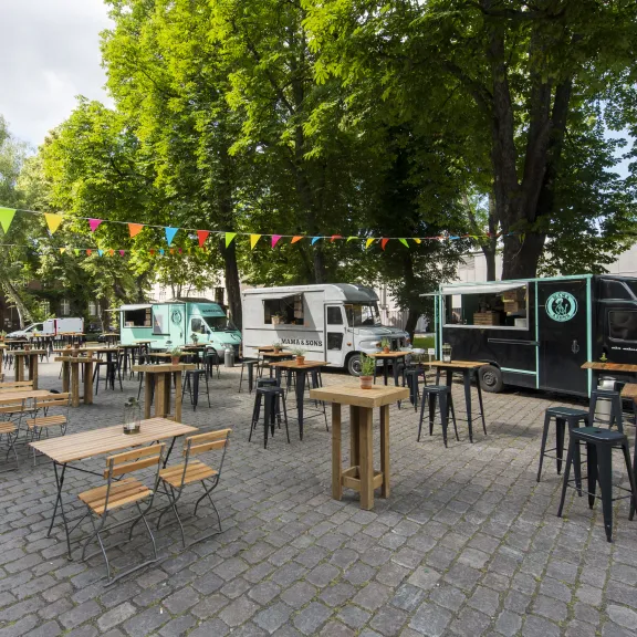 Leere Sitzbänke und Tische vor Foodtrucks auf einem gepflasterten Platz mit bunten Wimpeln.