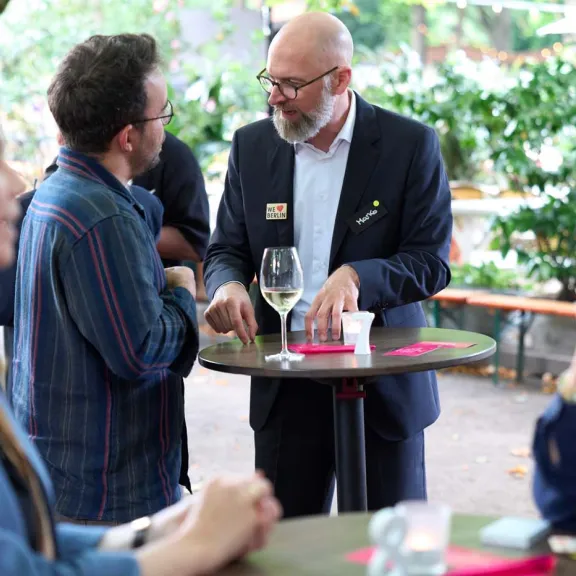 Menschen unterhalten sich an Stehtischen im Freien, mit Getränken und Snacks.