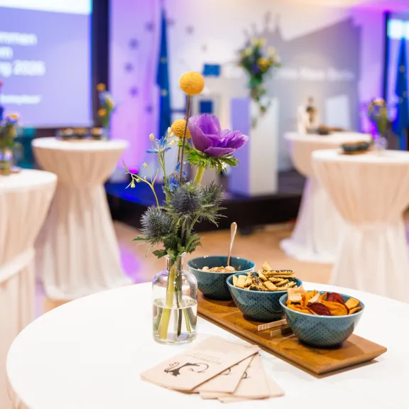 Hochtisch mit Snacks und Blumenvase, im Hintergrund eine Bühne bei einer Veranstaltung.