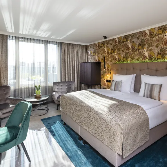 Modernes Hotelzimmer mit großem Bett, zwei Sesseln, Schreibtisch und bodentiefem Fenster.