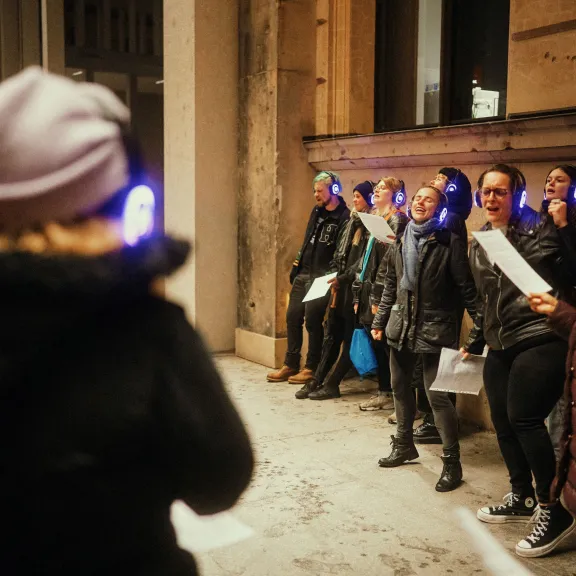 Gruppe mit leuchtenden Kopfhörern steht im urbanen Außenraum und führt eine gemeinsame Gesangs- oder Teamaufgabe während einer Silent Disco Tour durch.