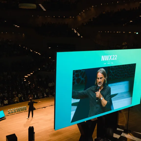 Großer Vortragssaal mit Sprecher auf Bühne und Präsentation auf Leinwand.