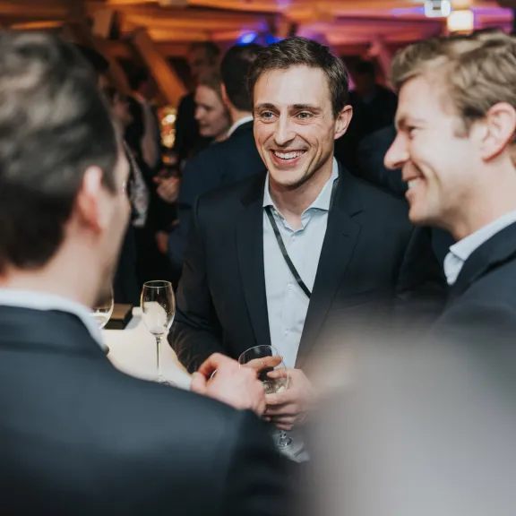 Drei Männer in Anzügen unterhalten sich lachend auf einer Veranstaltung.