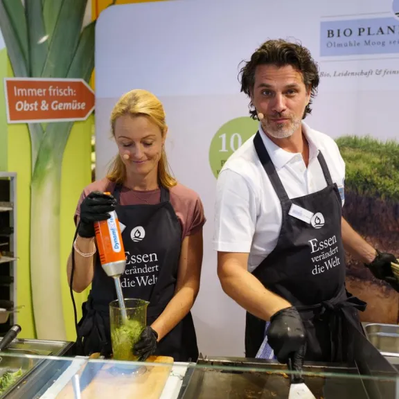 Zwei Personen in Kochschürzen bereiten Speisen an einem Messestand zu.