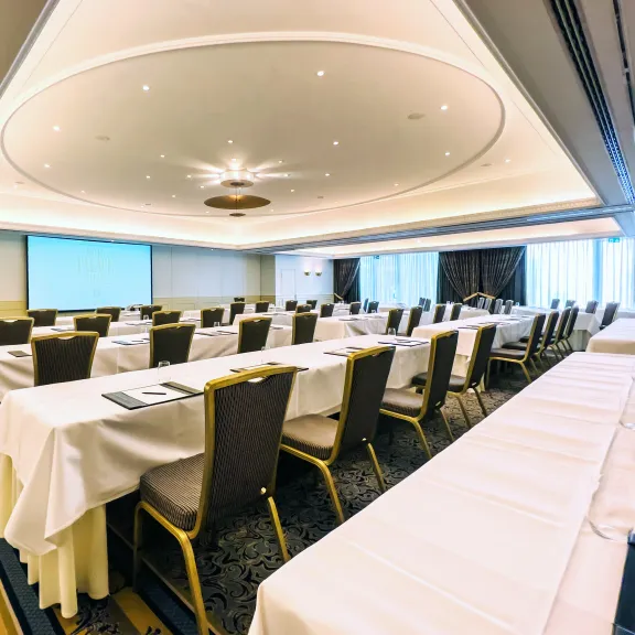 Tagungsraum mit langen, gedeckten Tischen und Stühlen, großer Bildschirm vorne