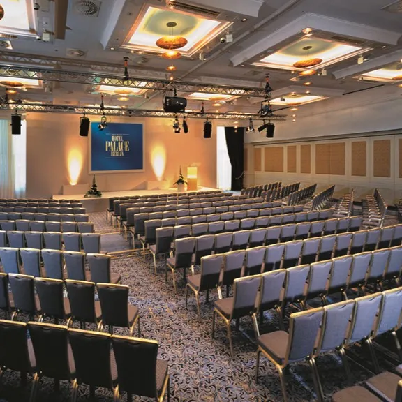 Moderner Konferenzraum im Hotel Palace Berlin mit flexiblen Sitzgelegenheiten, eleganten grauen Stühlen, einer Bühne mit Podium und Leinwand in einem gut beleuchteten, mit Teppich ausgelegten Raum.