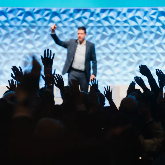 Mann auf Bühne spricht, Publikum hebt Hände, blaue Wand im Hintergrund.