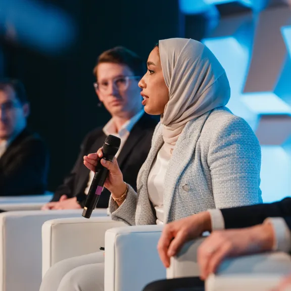 Frau im Hijab spricht mit Mikrofon auf Podium, drei weitere Personen hören zu.