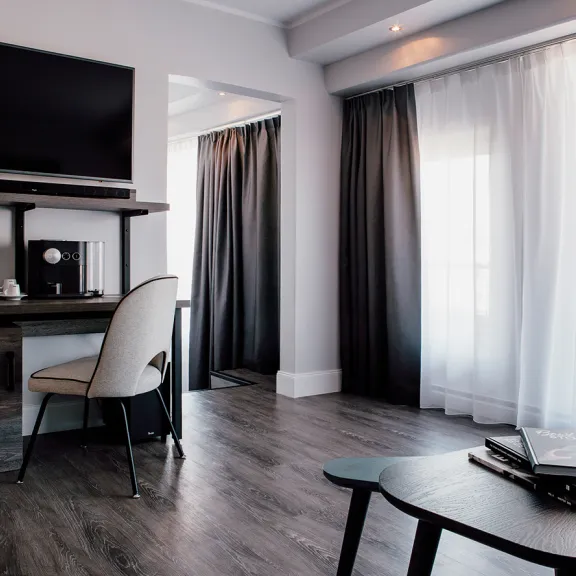 Modernes Hotelzimmer mit Schreibtisch, Stuhl, Fernseher und großen Fenstern mit Vorhängen.