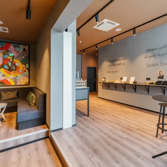 Modernes, helles Café mit Sitzecke, buntem Wandbild und Hochtischen auf Holzfußboden.