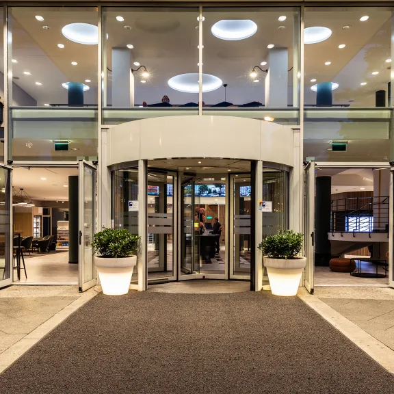 Hoteleingang mit Glasdach, Drehtür und zwei bepflanzten Kübeln bei Nacht.