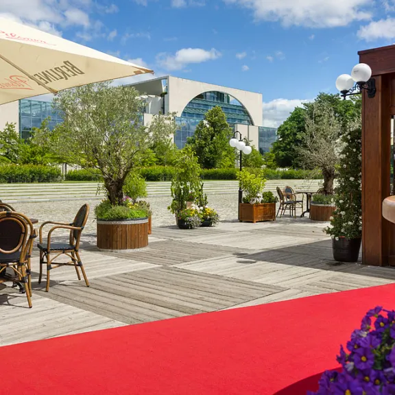 Sonnige Terrasse mit roten Teppich, Sitzbereich, Blumen und grüner Umgebung, Kanzleramt im Hintergrund.